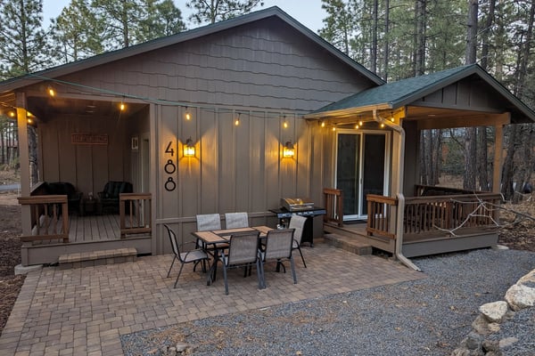 Red log cabin with turquoise roof and wooden deck surrounded by tall trees, grill in foreground